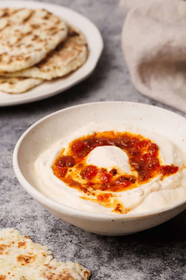 A white bowl filled with harissa yogurt next to a tea towel and flatbread.