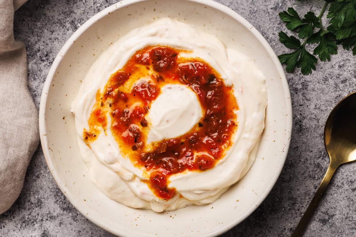 A white bowl filled with harissa yogurt next to a golden spoon, tea towel, and fresh parsley.