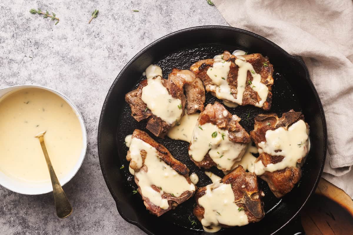 Lamb loin chops in a cast iron skillet next to a saucepan with a creamy sauce.