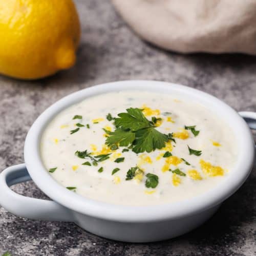 Lemon yogurt sauce in a small white enamel dish next to a lemon and natural linen tea towel.