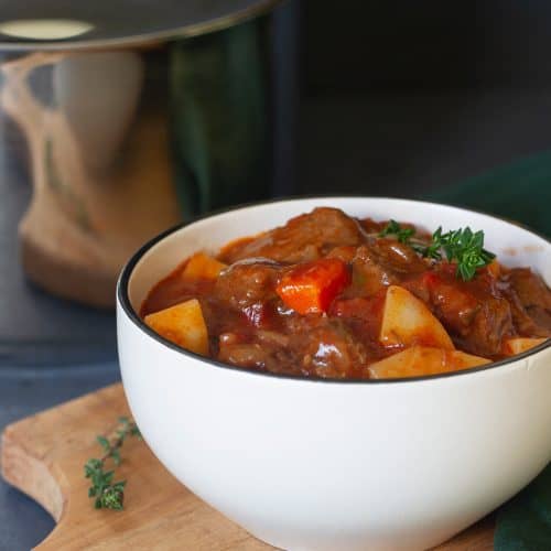 Beef stew in a small white bowl next to a large pot.
