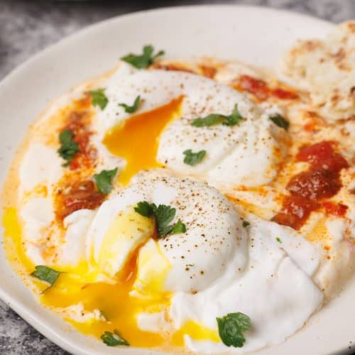 Turkish eggs with harissa on a white plate next to flatbread.