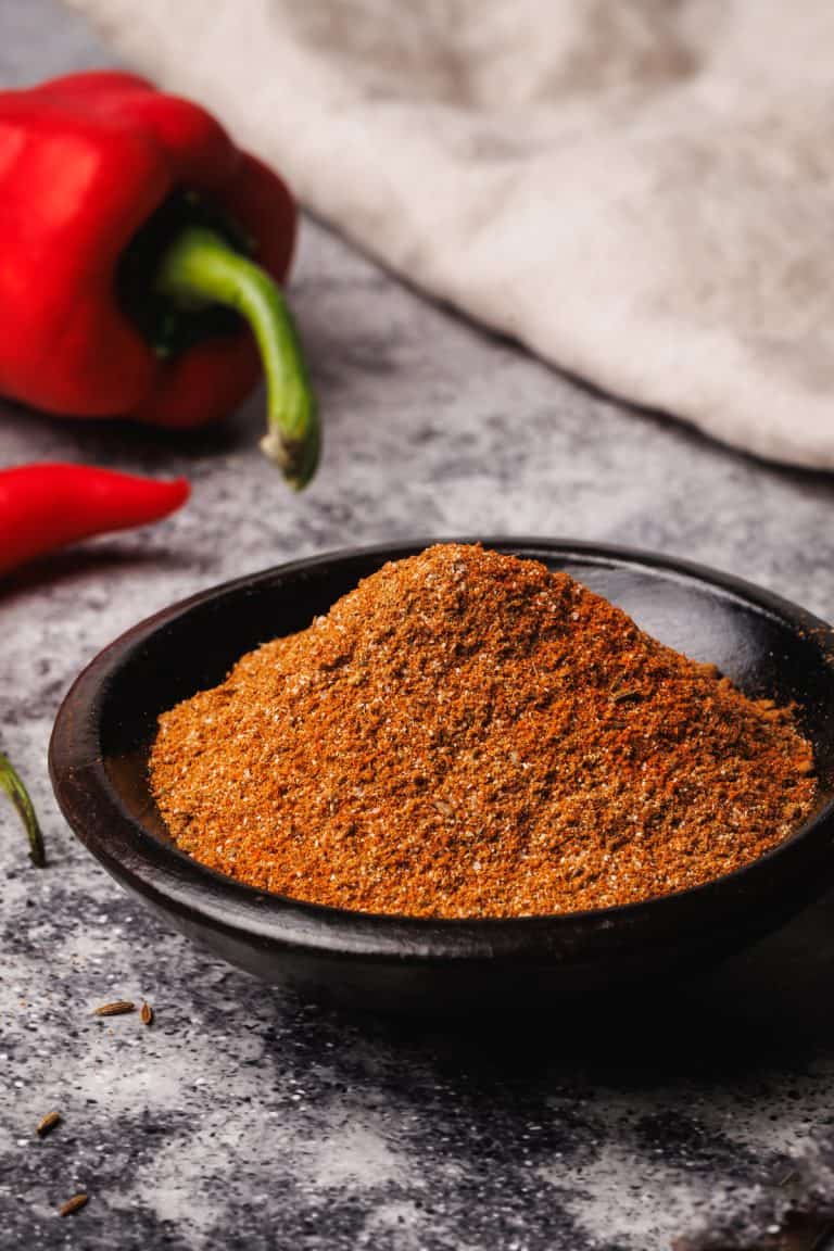 Harissa powder in a wooden bowl next to a golden spoon, peppers, and a tea towel.