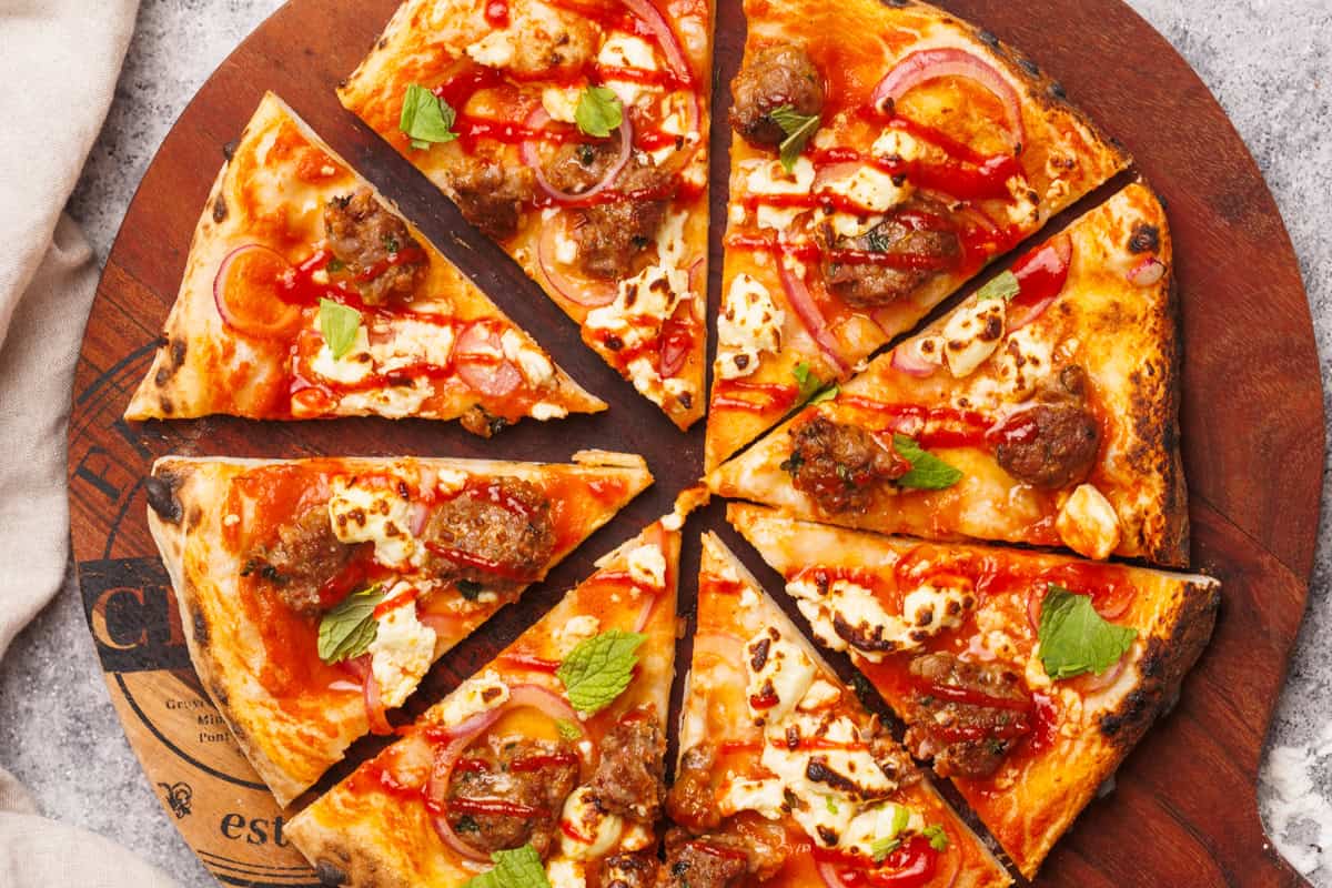 Cut lamb sausage pizza on a round wooden board.