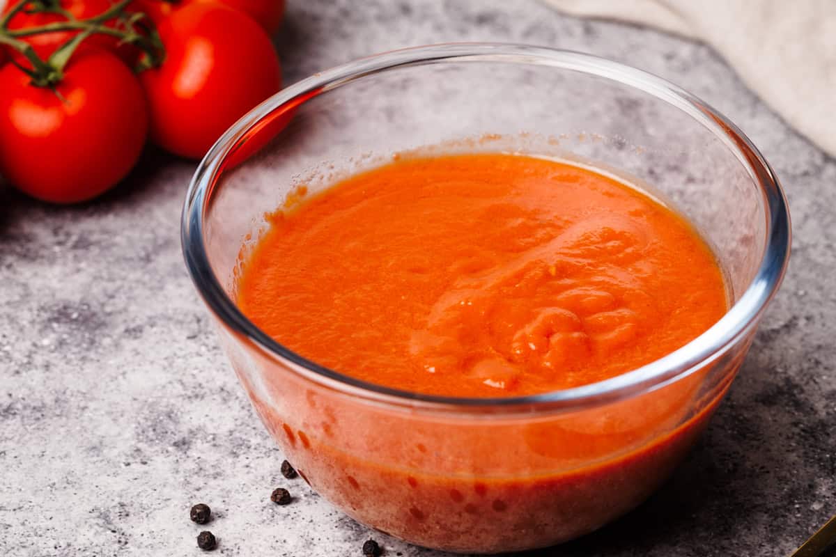 Pizza sauce in a glass bowl next to fresh tomatoes, a napkin, and black pepper.