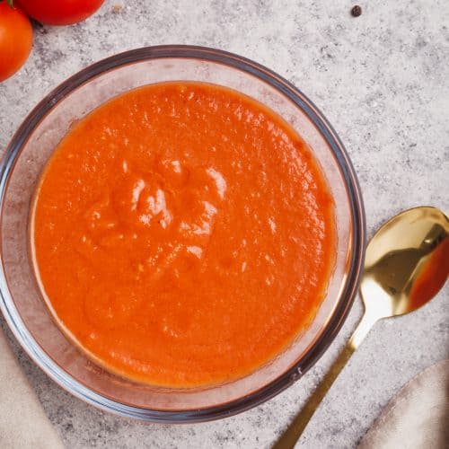 Pizza sauce in a glass bowl next to fresh tomatoes, a napkin, and black pepper.