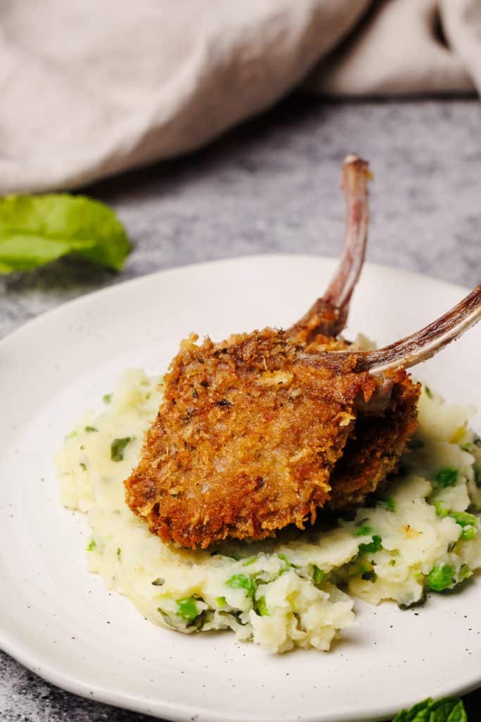Two crumbed lamb chops in some minted pea and potato mash on a white plate.