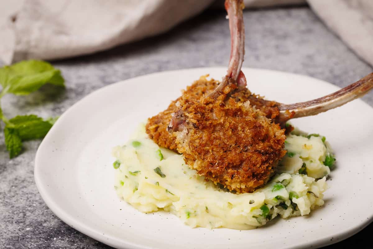 Two crumbed lamb chops in some minted pea and potato mash on a white plate.