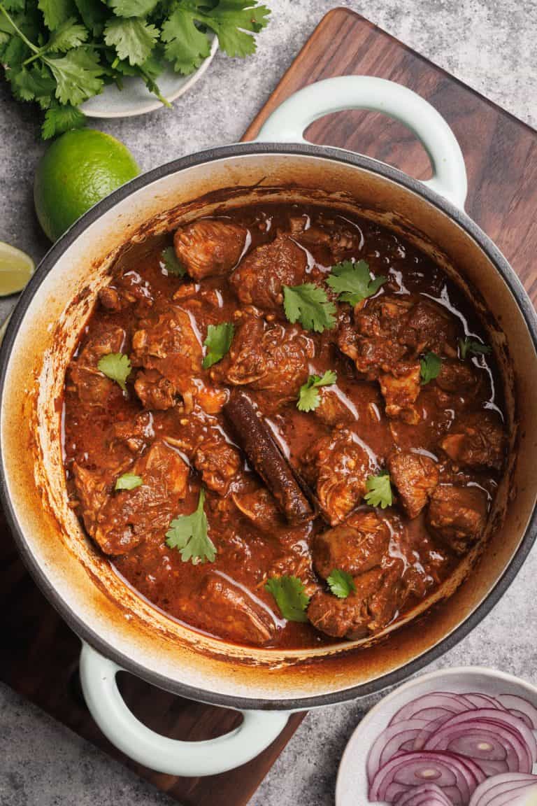 Lamb birria in a Dutch oven next lime, red onion, and coriander.