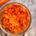Finished shaved carrot salad in a glass bowl next to carrots and a linen napkin.