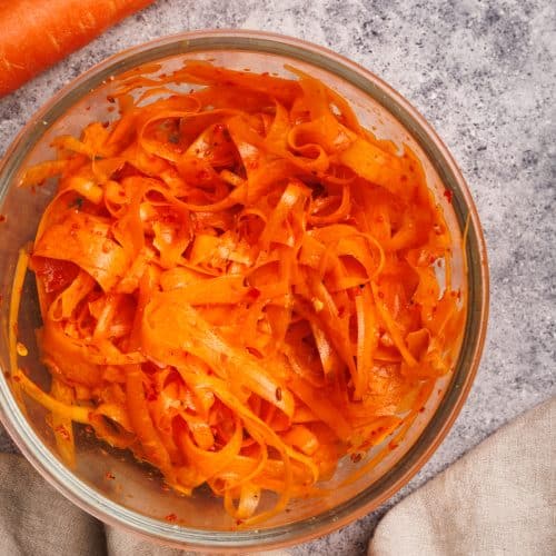 Finished shaved carrot salad in a glass bowl next to carrots and a linen napkin.