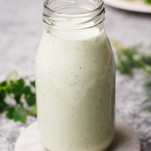 A small glass bottle filled with Greek yogurt ranch dressing on a marble coaster next to fresh herbs and a linen tea towel.