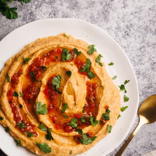 Harissa hummus on a white plate next to fresh herbs and a linen napkin.