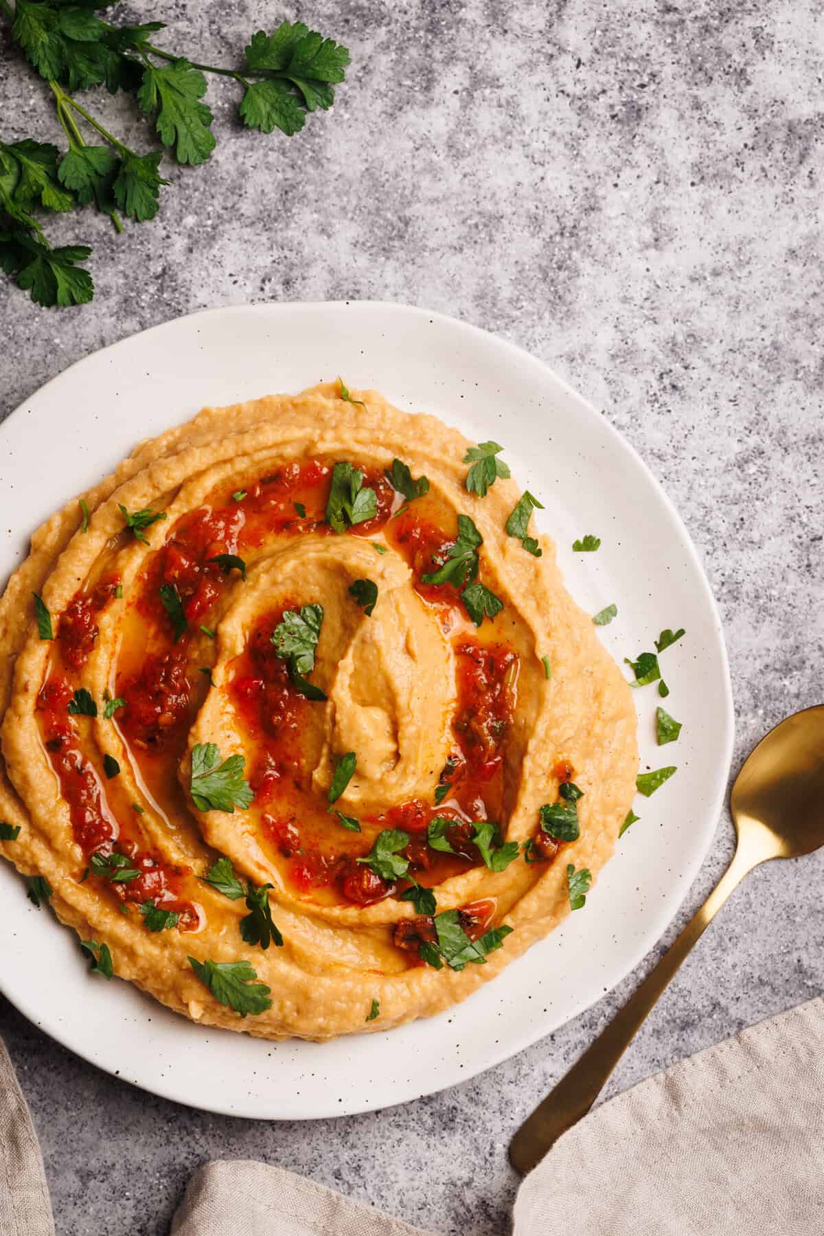 Harissa hummus on a white plate next to fresh herbs and a linen napkin.