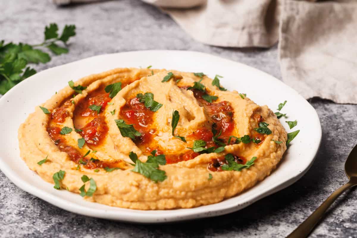 Harissa hummus on a white plate next to fresh herbs and a linen napkin.