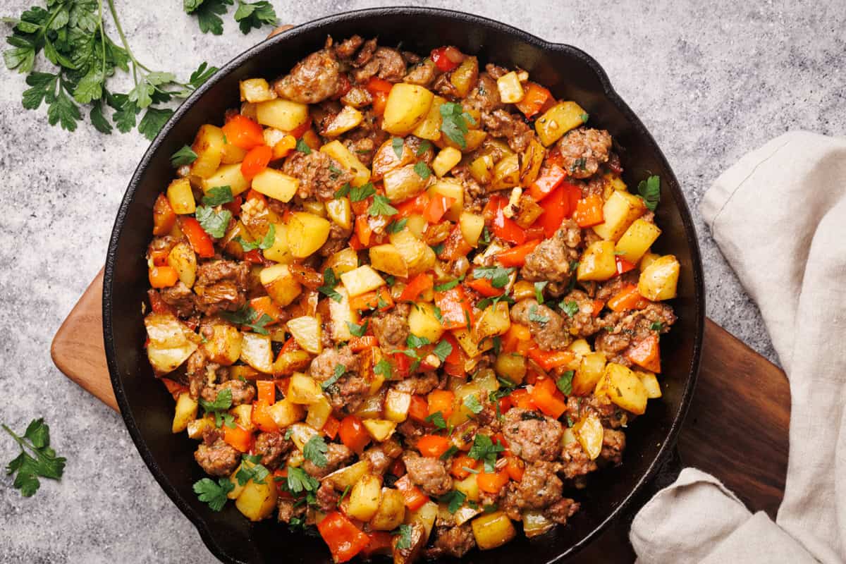 Sausage and potato hash in a cast iron pan next to a linen towel and fresh herbs.