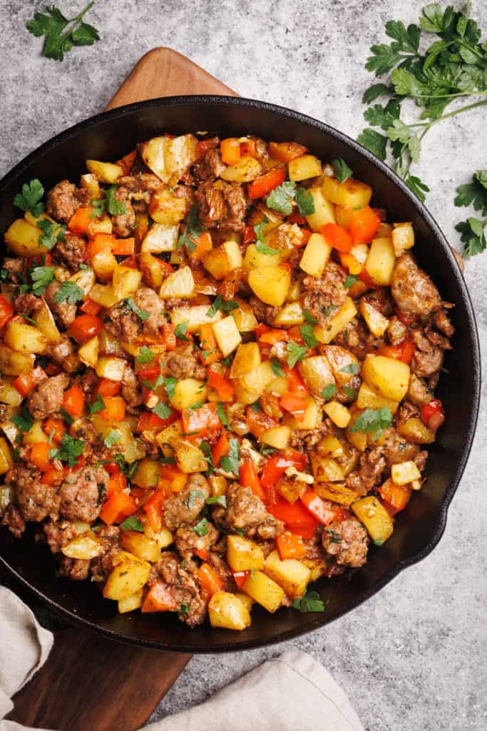 Sausage and potato hash in a cast iron pan next to a linen towel and fresh herbs.